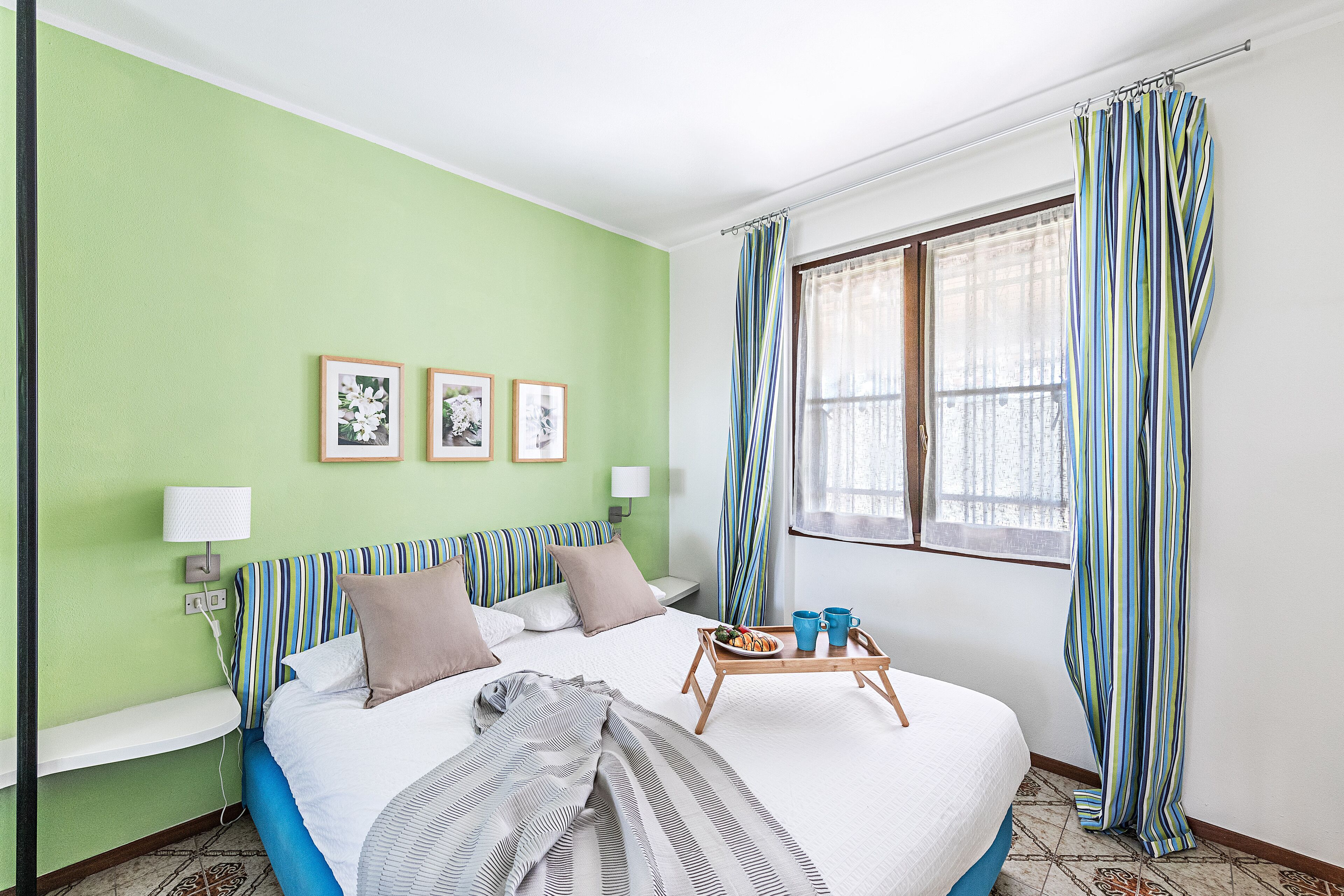 Hotellihuone, jossa on sänky, yöpöytä, ikkuna verhoilla ja seinä, jossa on kolme kehystettyä kuvaa.