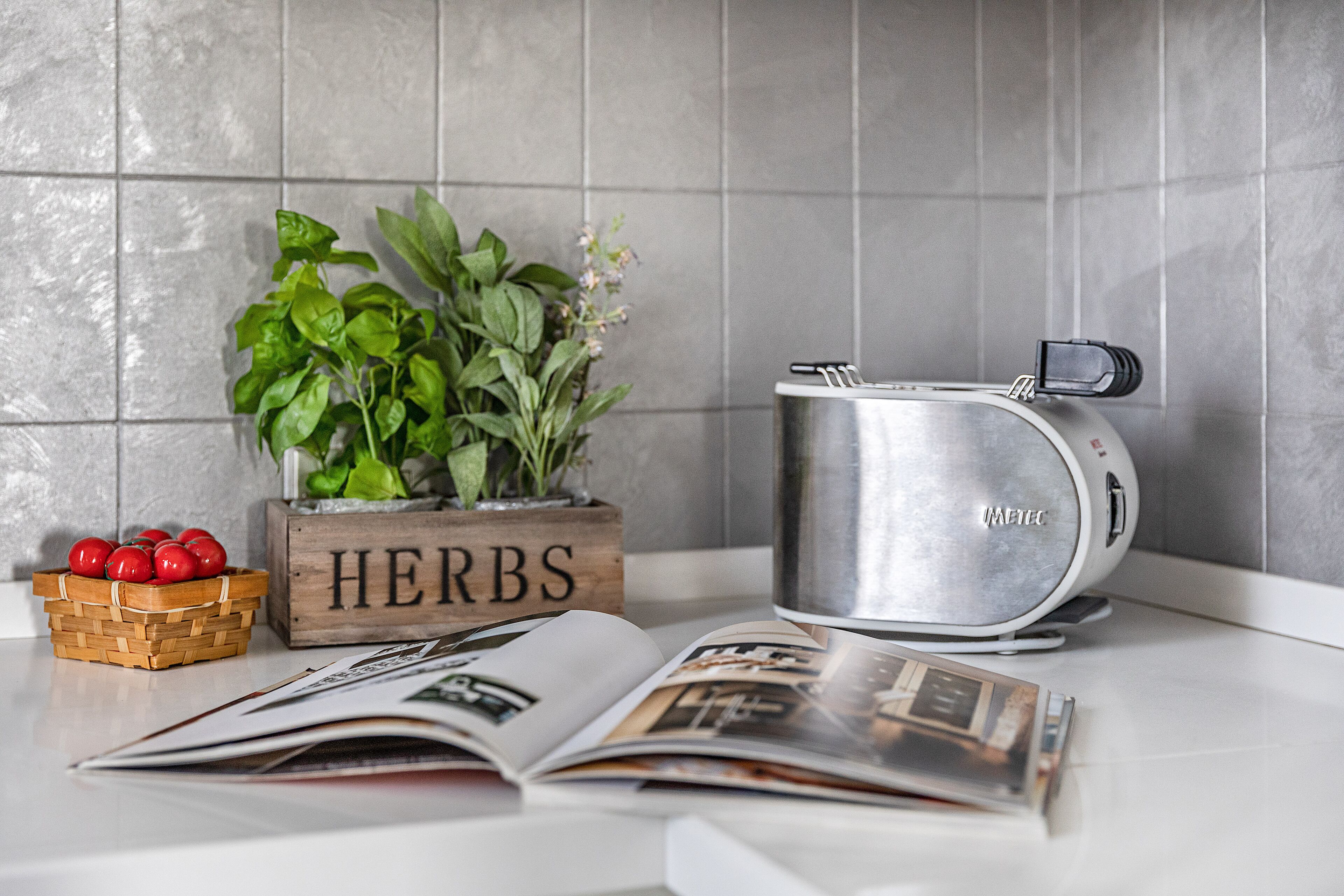 A kitchen counter with a stainless steel pot, a wooden block with the word "HERBS", a basket of red tomatoes, and an open magazine.