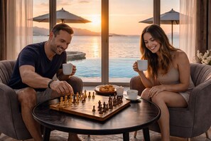 A couple playing chess in a room with a view of the sea at sunset.