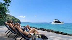 Ein Paar entspannt auf Liegestühlen am Strand, im Hintergrund liegt eine Yacht.