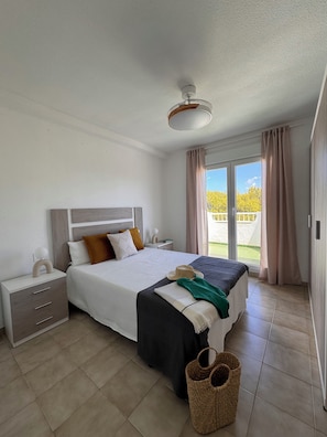 Un dormitorio con cama, mesitas de noche, ventilador de techo, ventana con cortinas y vistas a zonas verdes.