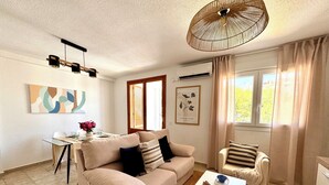 Una sala de estar con un sofá color beige, una mesa de centro blanca, un sillón a rayas, una mesa de comedor con sillas, una lámpara colgante y una ventana con cortinas.