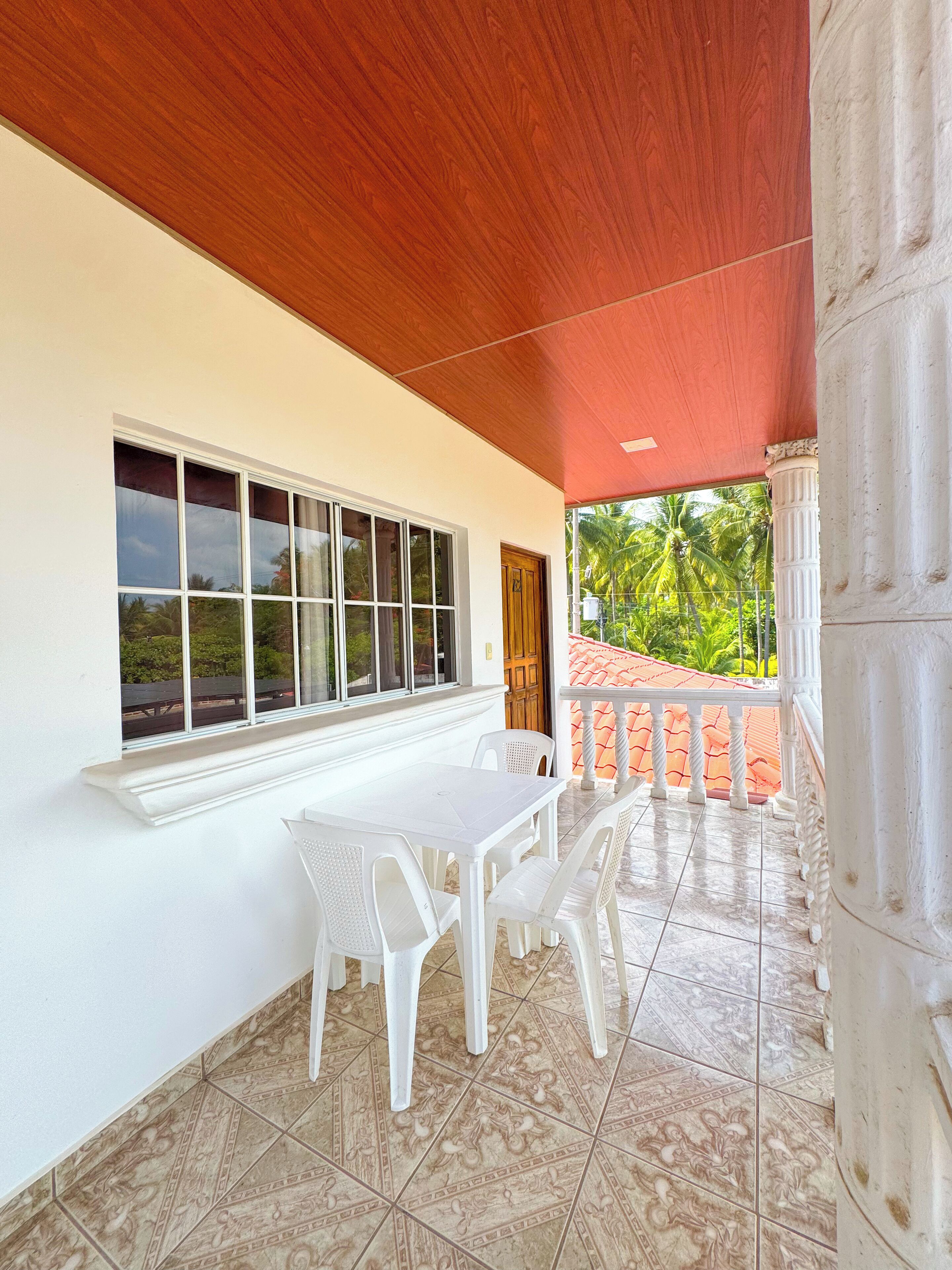 Un patio cubierto con una mesa y sillas blancas, una ventana con vista a follaje y un suelo de baldosas.