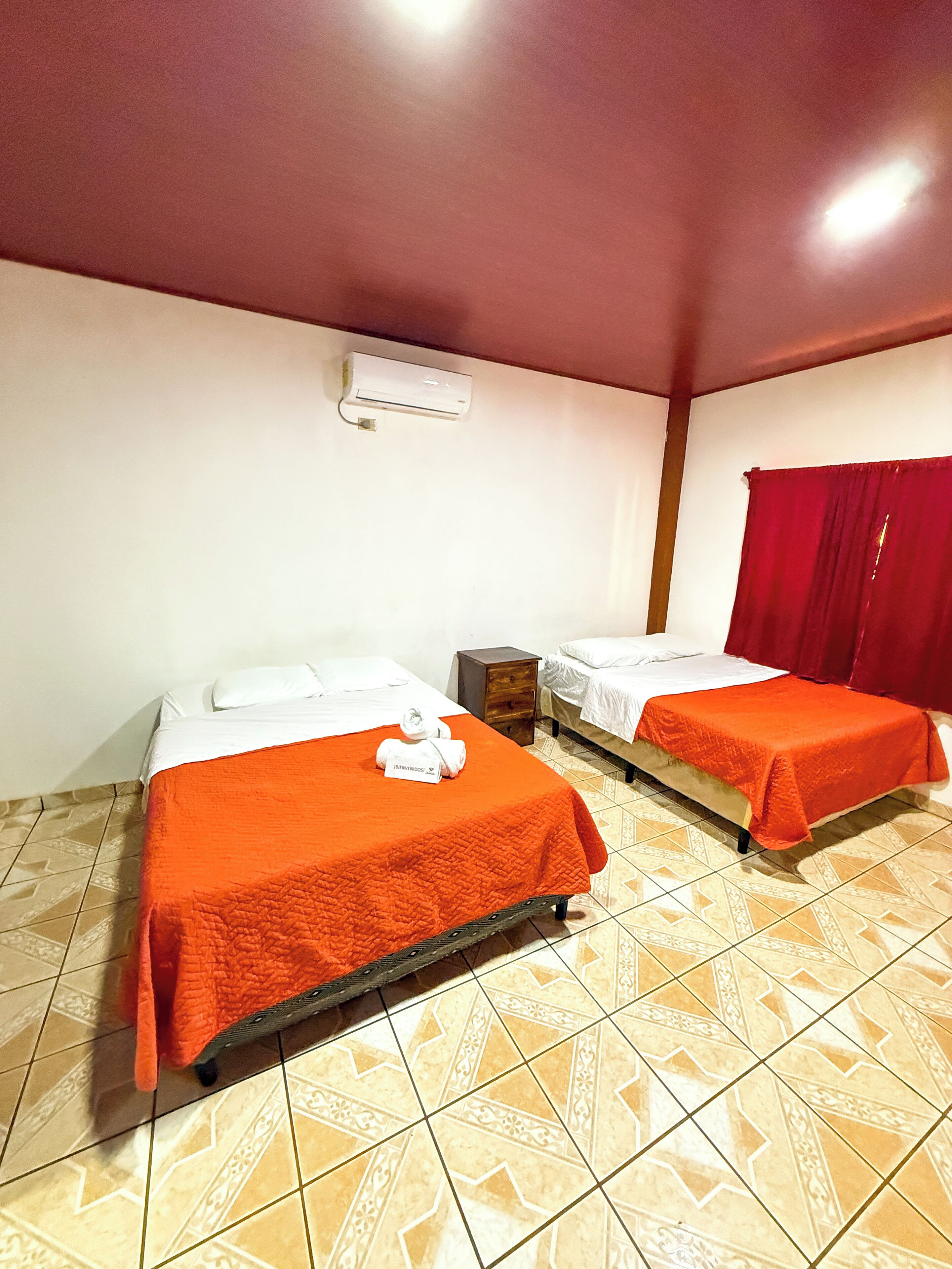 A room with two beds, each with an orange bedspread and white pillows. There is a small wooden nightstand between the beds. The room has patterned tile flooring and a red curtain on the window.