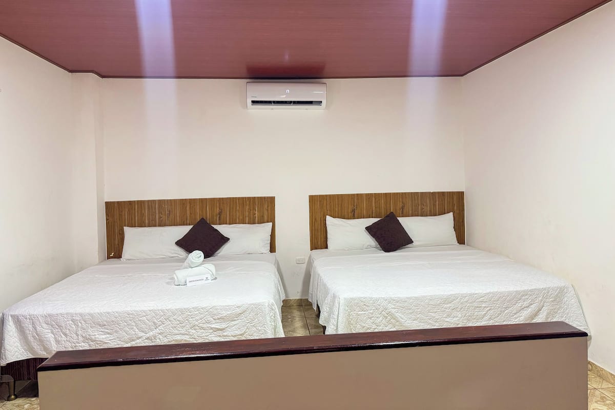 A hotel room with two single beds, each with a wooden headboard and white bedding. There is a small air conditioning unit on the wall above the beds.