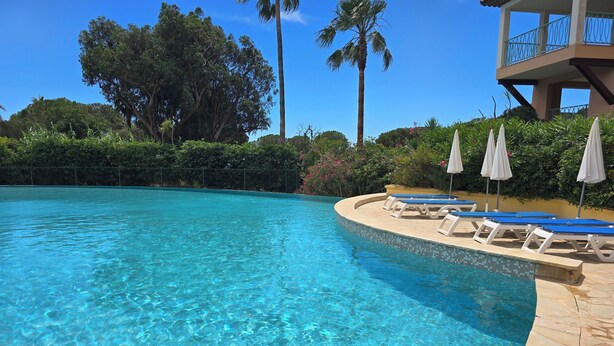 A clear blue pool with lounge chairs and umbrellas, surrounded by lush greenery and a building with a balcony. - Les rives des Issambres 1201 (Roquebrune-sur-Argens)