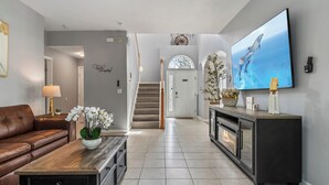 A modern living room with a brown leather sofa, a wooden coffee table with white flowers, a fireplace, and a large TV on the wall. - Luxury relaxing stay. A brand new remodel villa near Disney and Legoland  (Haines City)