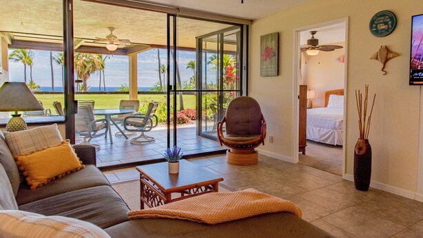 A spacious living area with a sofa, a coffee table, and a chair. There is a sliding glass door leading to an outdoor patio with a table and chairs. A ceiling fan is visible, and a bed is seen in the adjacent room. - Ocean & Golf Views | Remodeled Kona Villa w/ AC (Kailua-kona)
