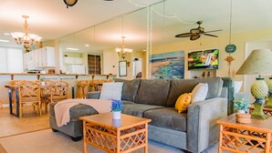 A living room with a grey sofa, wooden coffee table, and wicker chairs. There is a TV mounted on the wall and a ceiling fan. - Ocean & Golf Views | Remodeled Kona Villa w/ AC (Kailua-kona)