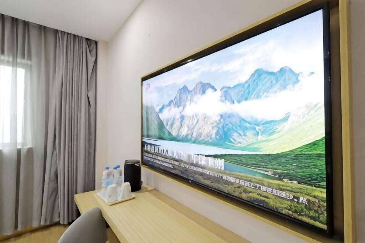 Quarto de hotel com uma televisão grande na parede exibindo uma paisagem montanhosa, uma mesa de madeira com um alto-falante e água engarrafada, e uma janela com cortinas.