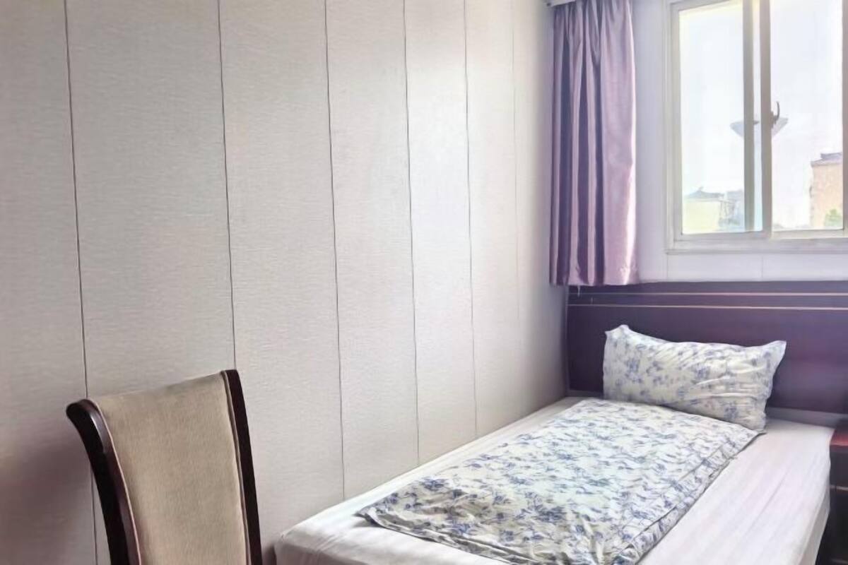 Uma cama de solteiro com edredom floral, uma cadeira de madeira e uma janela com cortinas.