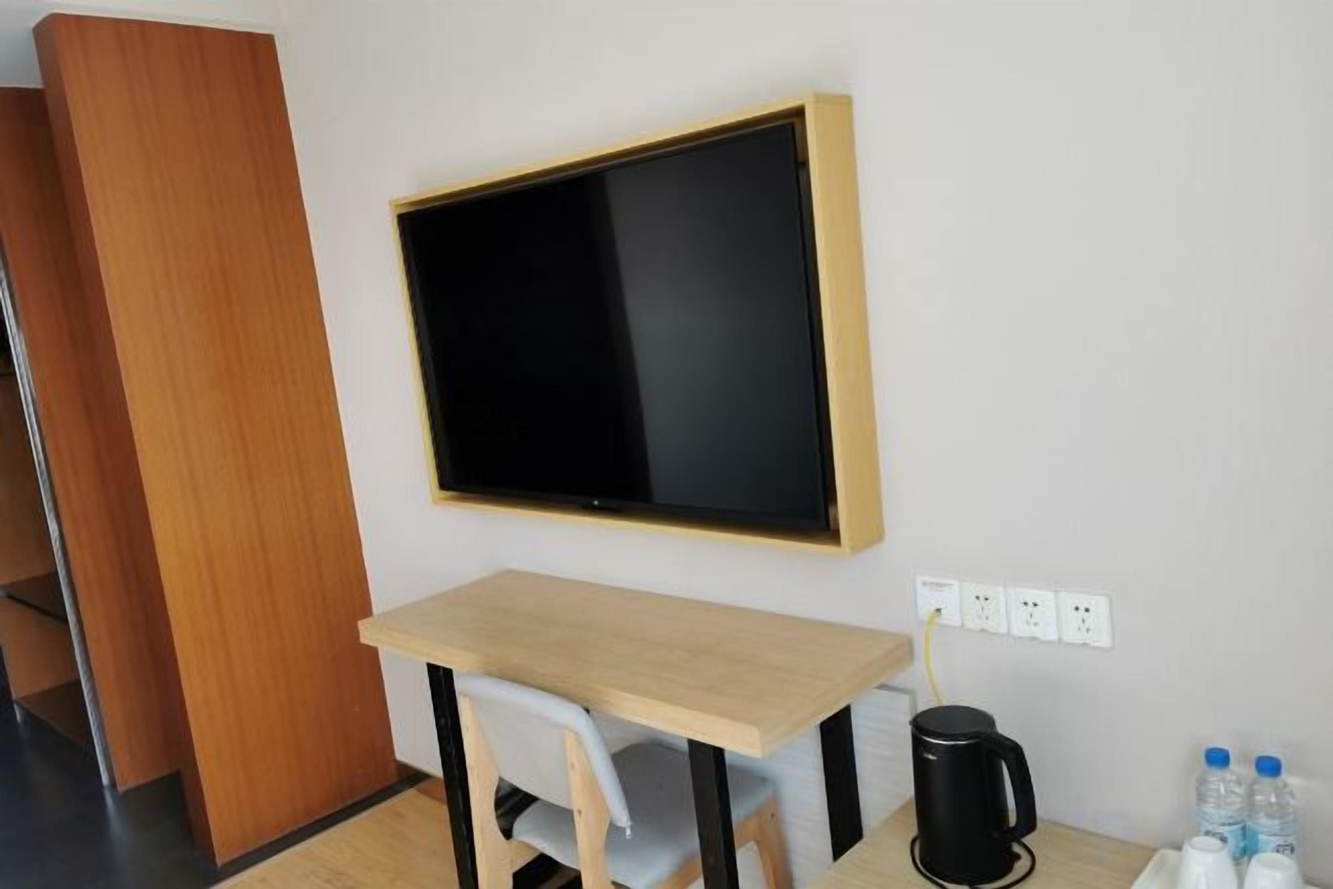 Quarto com escrivaninha de madeira, cadeira preta, televisão de tela plana na parede, cafeteira e tomada.