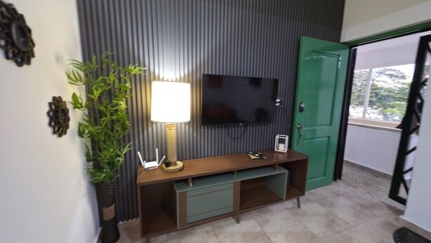 Uma sala de estar moderna com uma TV de tela plana na parede, um móvel de entretenimento de madeira com gavetas, uma porta verde e um vaso com planta.