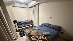 A small, clean room with a single bed, a second bed in the background, a standing fan, and a tiled floor. - A 4 Minutos da Basílica! Apartamento Para Famílias (Aparecida)