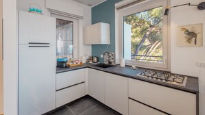 A modern kitchen with white cabinets, a gas stove, a window with a view of trees, and a wall-mounted light fixture. - Sky & Sea Apartment by Wonderful Italy (Ospedaletti)