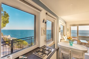 Une cuisine moderne équipée d’une cuisinière à gaz, de grandes fenêtres donnant sur la mer et d’un coin repas avec une table et des chaises blanches.