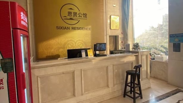 Un comptoir d’accueil avec une enseigne indiquant « SIXIAN RESIDENCE » et un écran d’ordinateur. Un distributeur automatique rouge se trouve à gauche, et une fenêtre offre une vue sur les bâtiments à l’extérieur.