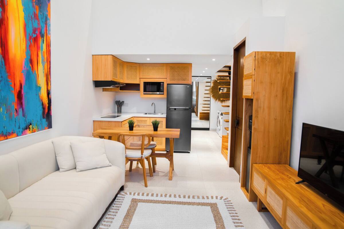 Un salon moderne comprenant un canapé, une table à manger avec des chaises, un meuble pour la télévision et un coin cuisine avec des meubles et des appareils électroménagers en bois.