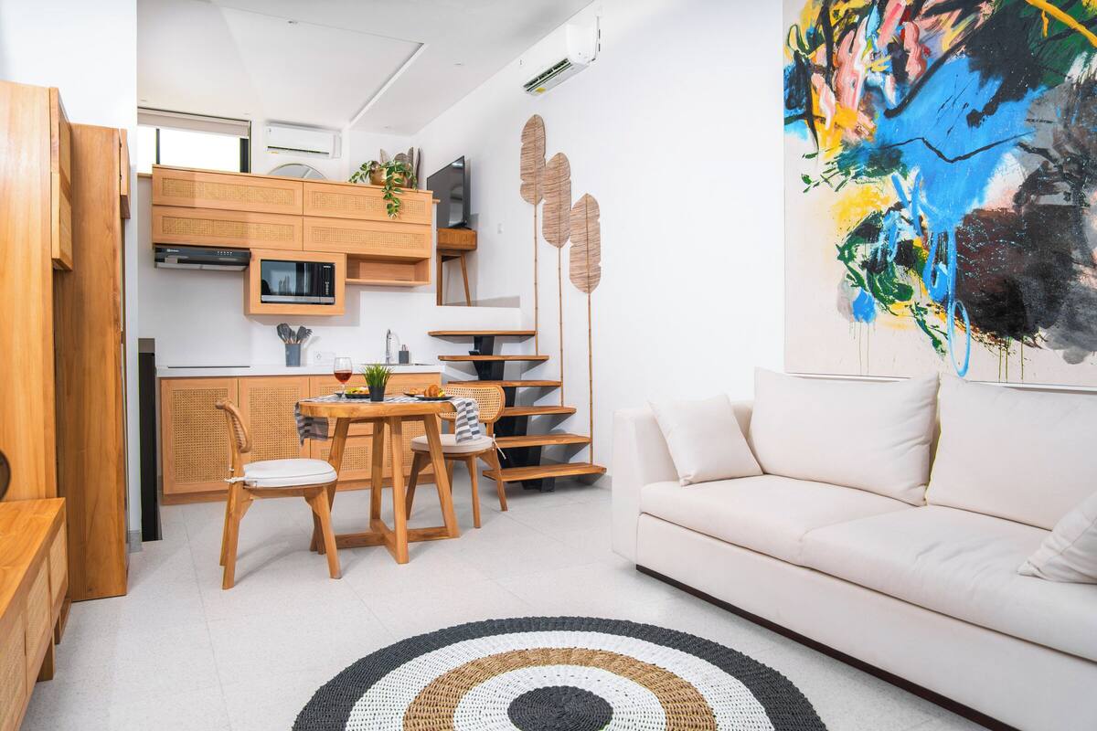 Une cuisine moderne avec des meubles en bois, un coin repas avec une table et des chaises, et un salon avec un canapé et un grand tableau abstrait.