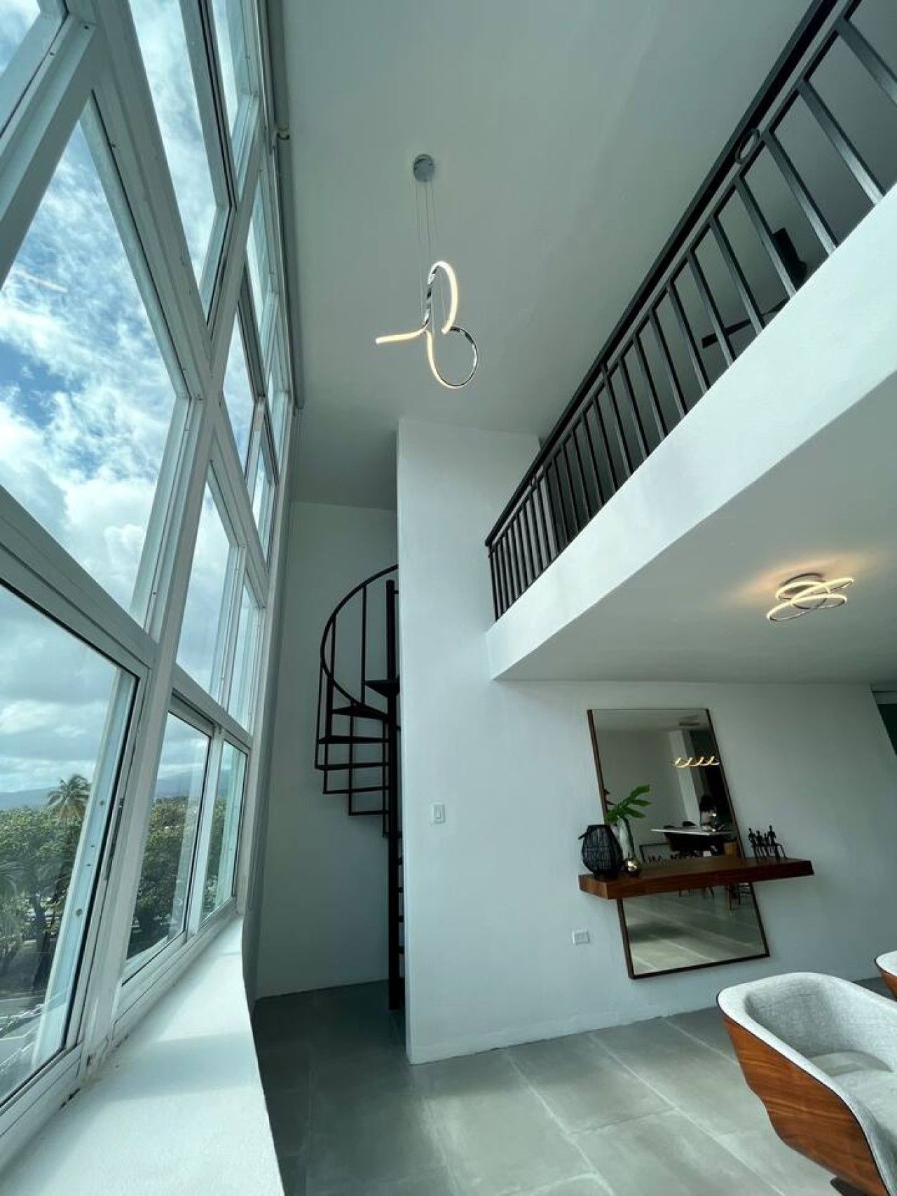 Amplo interior com grandes janelas, escadaria em espiral e luminária moderna.