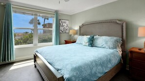 Une chambre à coucher comprenant un lit, une table de chevet, un ventilateur de plafond, une fenêtre avec des rideaux et un mur orné d’un cadre contenant une citation.