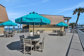 An outdoor patio area with tables and chairs, shaded by large umbrellas, and a view of the ocean. - Coquina A104 Ground floor walk out unit. Direct Ocean View! (Saint Augustine Beach Florida)