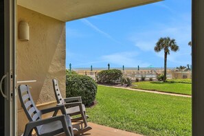 A balcony with two chairs, a view of a well-maintained lawn, and a distant beach with palm trees. - Coquina A104 Ground floor walk out unit. Direct Ocean View! (Saint Augustine Beach Florida)