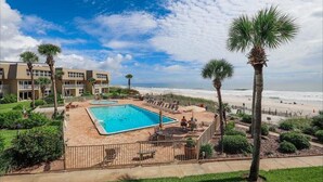Un espace piscine offrant une vue sur la plage, entouré de palmiers et bordé d’un immeuble doté de nombreux balcons.