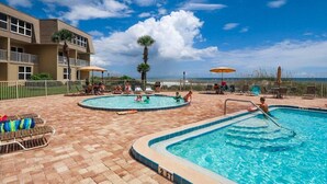 Zona de piscina amb dues piscines, gandules, ombrel·les i un edifici amb balcons.