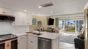 A modern kitchen with a built-in oven, microwave, and dishwasher, a dining area with a table and chairs, and a large sliding glass door leading to a balcony with a view of the ocean.