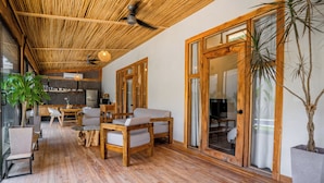A spacious room with a wooden floor, a dining area with chairs and a table, a kitchen area with a refrigerator, and a large window with a view. - Casa Heaven Santa Teresa (Cóbano)