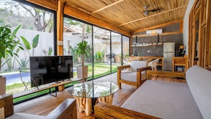 A modern living room with a glass coffee table, wooden furniture, a flat-screen TV, and large windows offering a view of the outdoors. - Casa Heaven Santa Teresa (Cóbano)