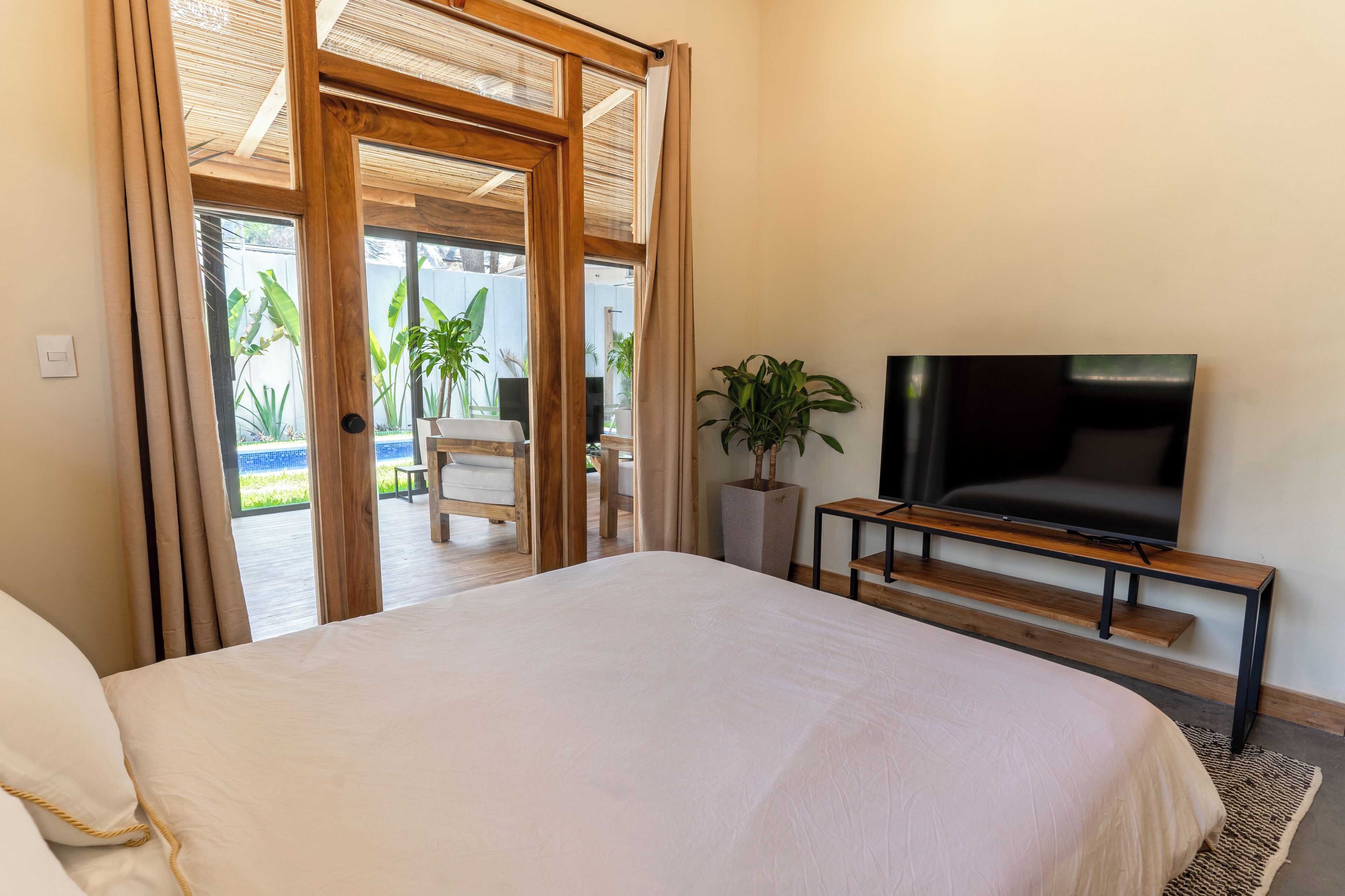 Une chambre avec un grand lit, un téléviseur posé sur un meuble en bois, une plante en pot et une porte-fenêtre coulissante donnant sur un espace extérieur aménagé avec une table et des chaises.