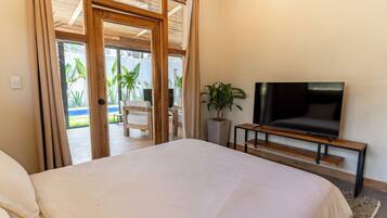 Une chambre avec un grand lit, un téléviseur posé sur un meuble en bois, une plante en pot et une porte-fenêtre coulissante donnant sur un espace extérieur aménagé avec une table et des chaises.