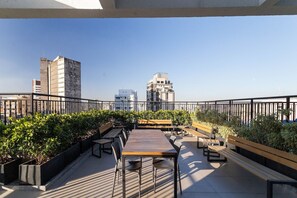 An outdoor terrace with a long wooden table and benches, surrounded by greenery and overlooking a cityscape with modern buildings. - My One Bela Vista (São Paulo)