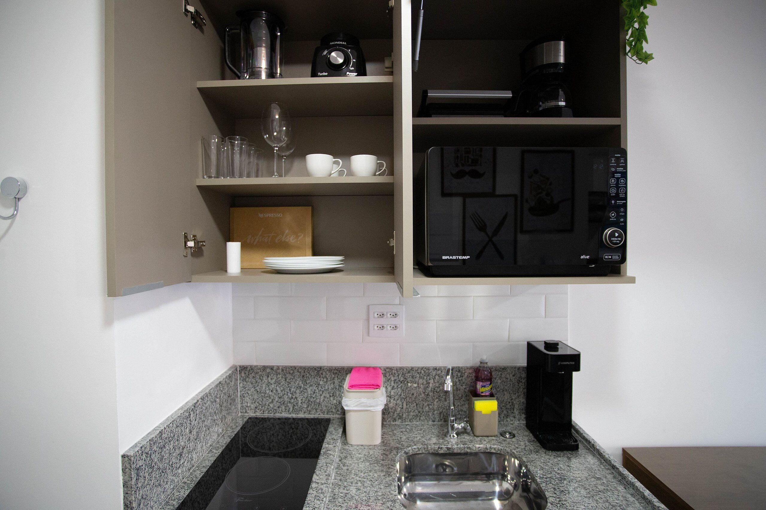Eine Küche mit Mikrowelle, Kaffeemaschine und Toaster. Es gibt Regale mit Gläsern und Tassen sowie ein Spülbecken mit Seifenspender.