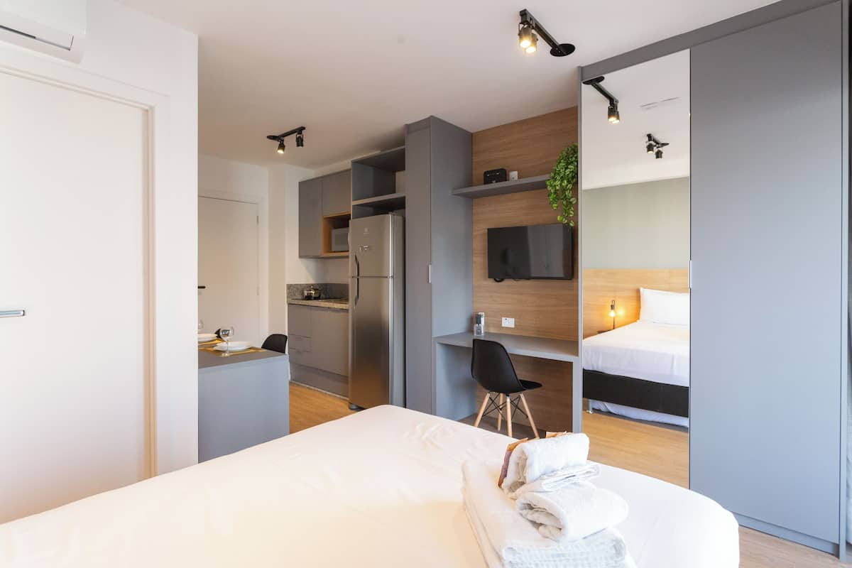 A modern hotel room with a bed, a desk, a kitchenette, and a TV.