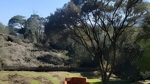 An outdoor seating area with two chairs and a table, surrounded by a natural landscape with trees and a hill in the background. - Red Barn Lindo Celeiro em Urubici - SC (Urubici)