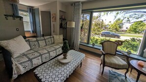A living room with a patterned sofa, a small table, a chair, and a large window with a view of the street. - Robin's Bluff- Classic 3-bedroom in beautiful Whitefish Bay, convenient location (Whitefish Bay)