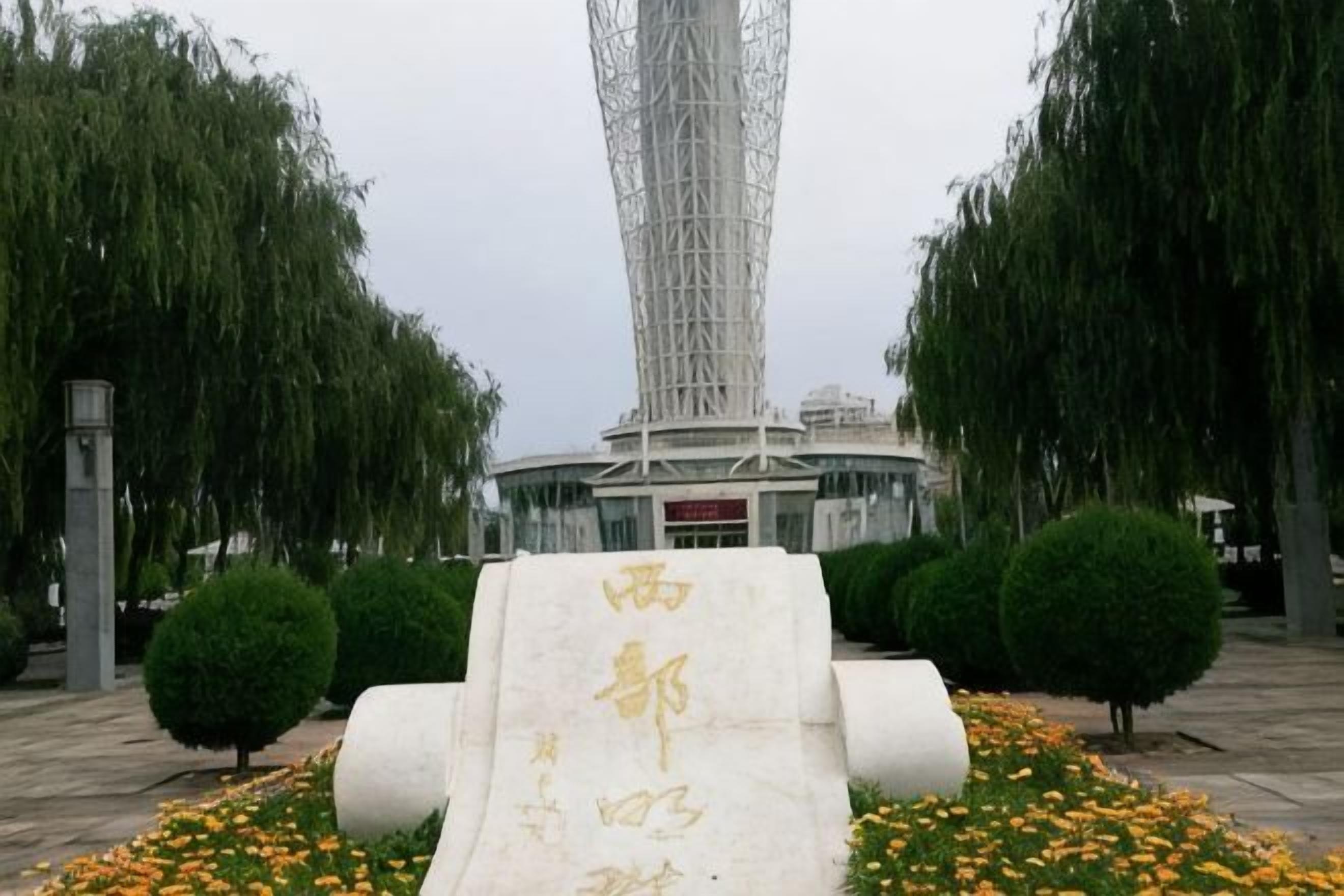 Um monumento com inscrições, rodeado por canteiros de flores e árvores, com uma estrutura alta ao fundo.