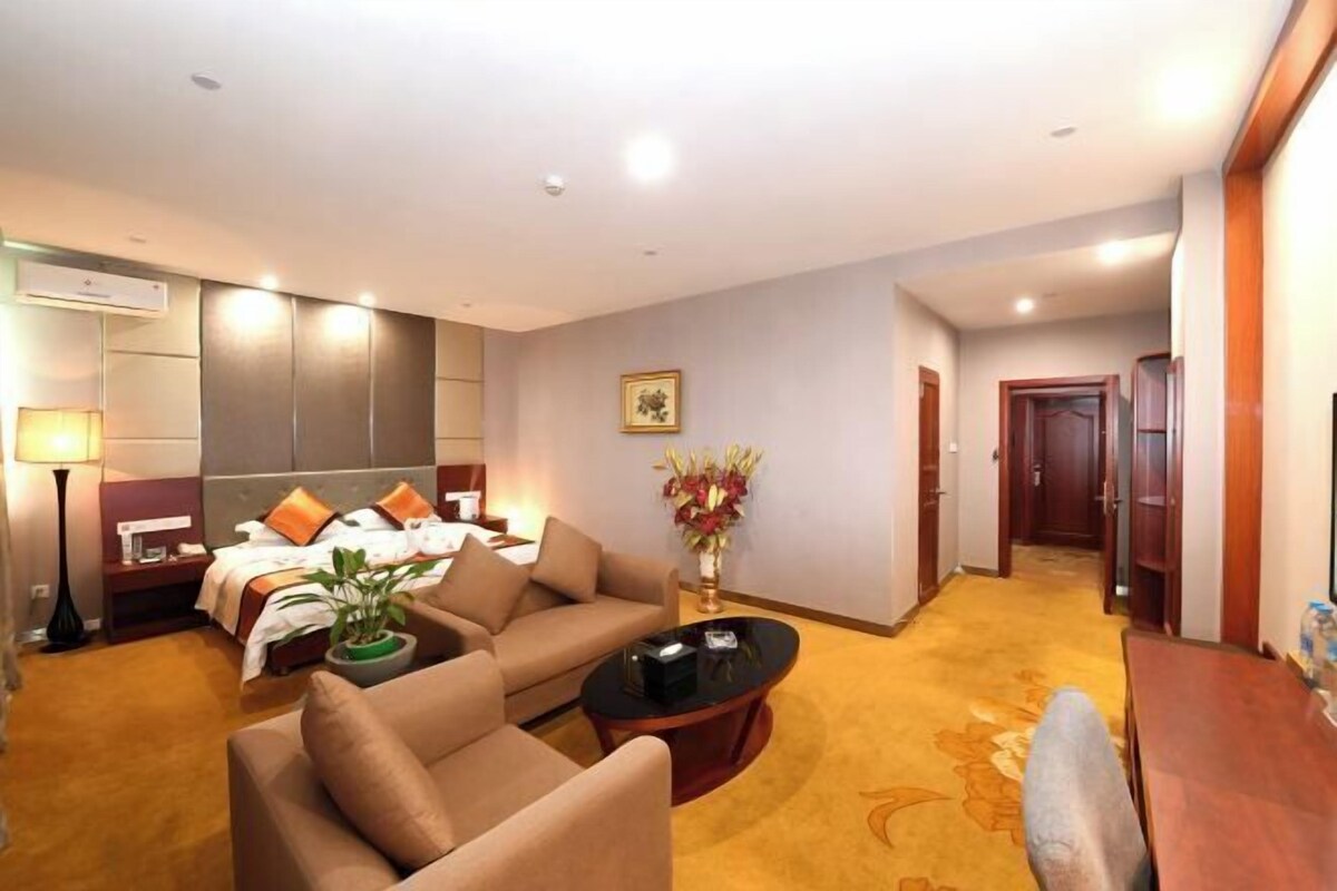 Habitación de hotel con cama, sofá, mesa de centro, lámpara, jarrón con flores y un pasillo que conduce a una puerta.