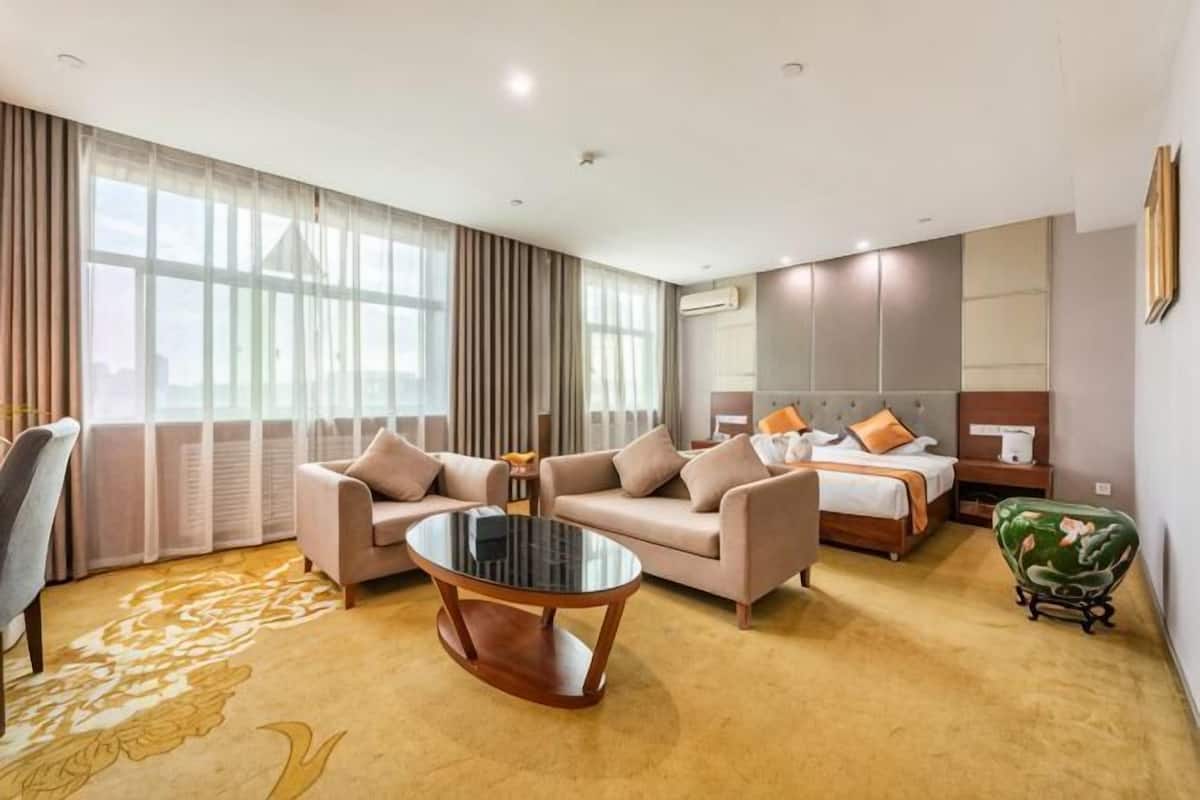 Une chambre d’hôtel avec un canapé, une table basse ronde, un lit avec des oreillers, une table de chevet et un fauteuil vert.