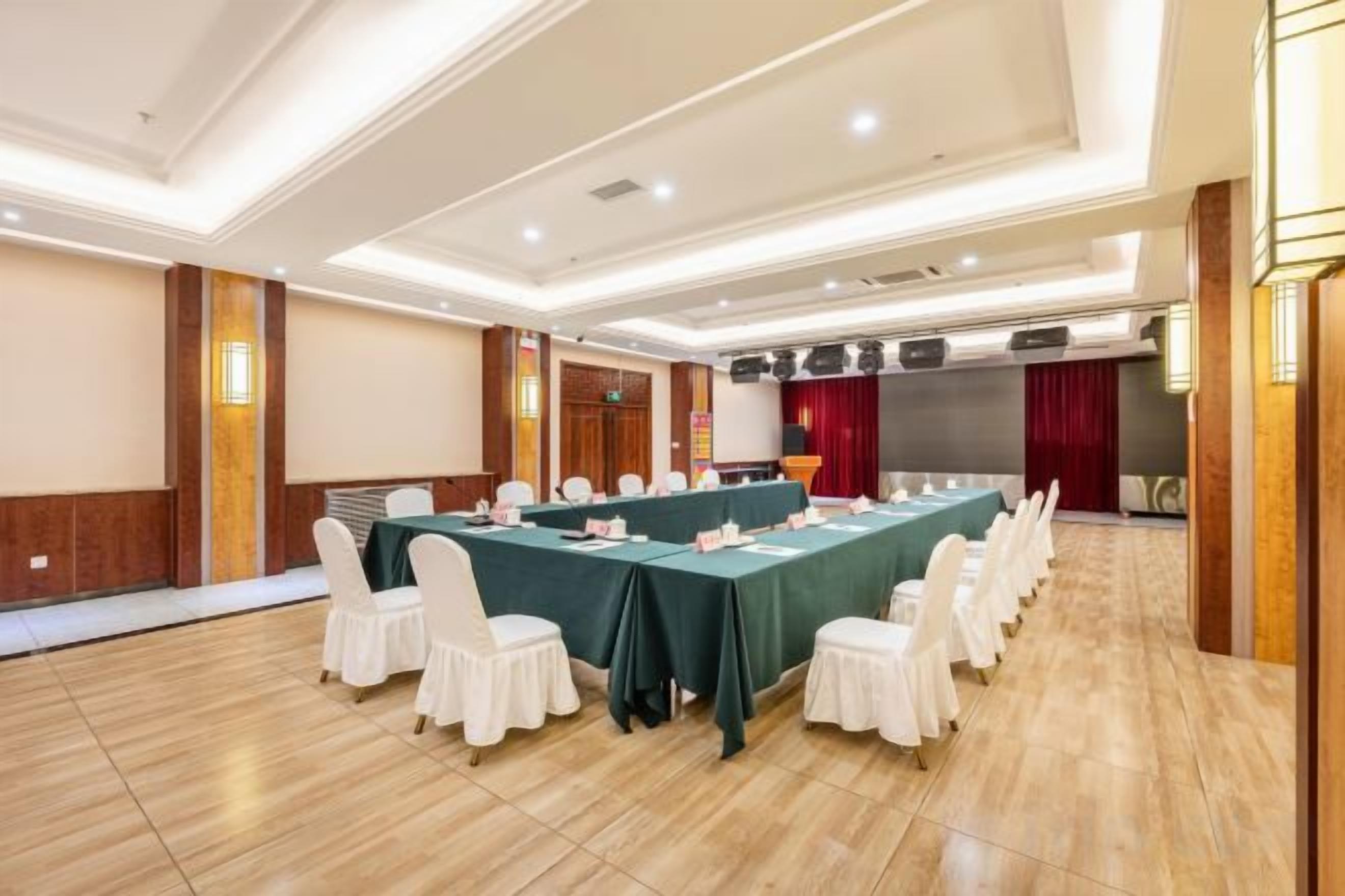 Un salón de conferencias con una larga mesa, sillas blancas y un escenario con una cortina roja.