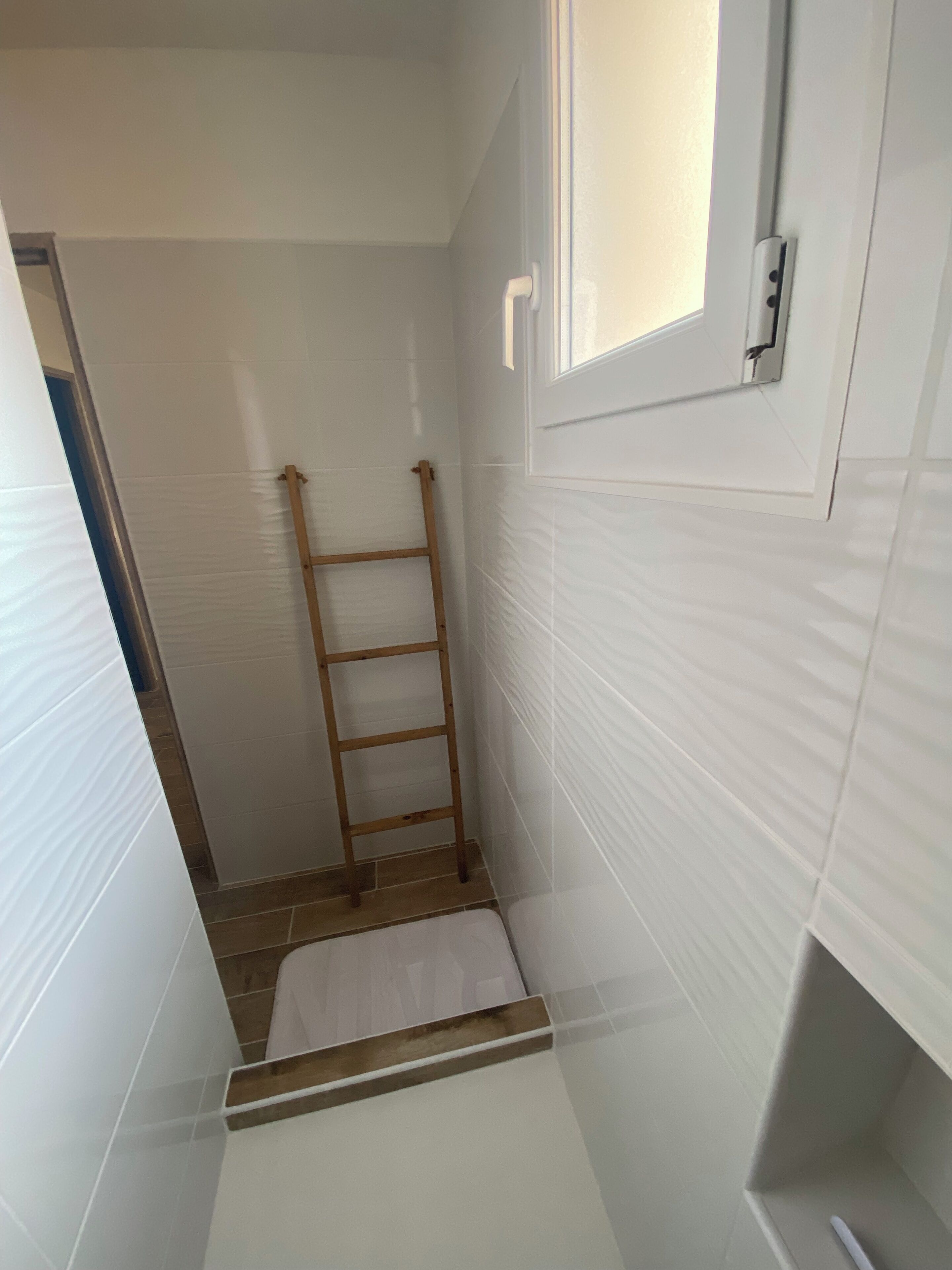 Un baño estrecho con paredes de azulejos blancos, una escalera de madera y un ventanal pequeño.