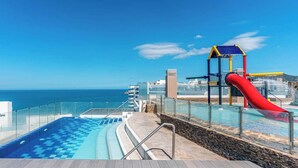 Un espace piscine avec un toboggan rouge, une structure de jeux bleue et jaune, et une vue dégagée sur l’océan.