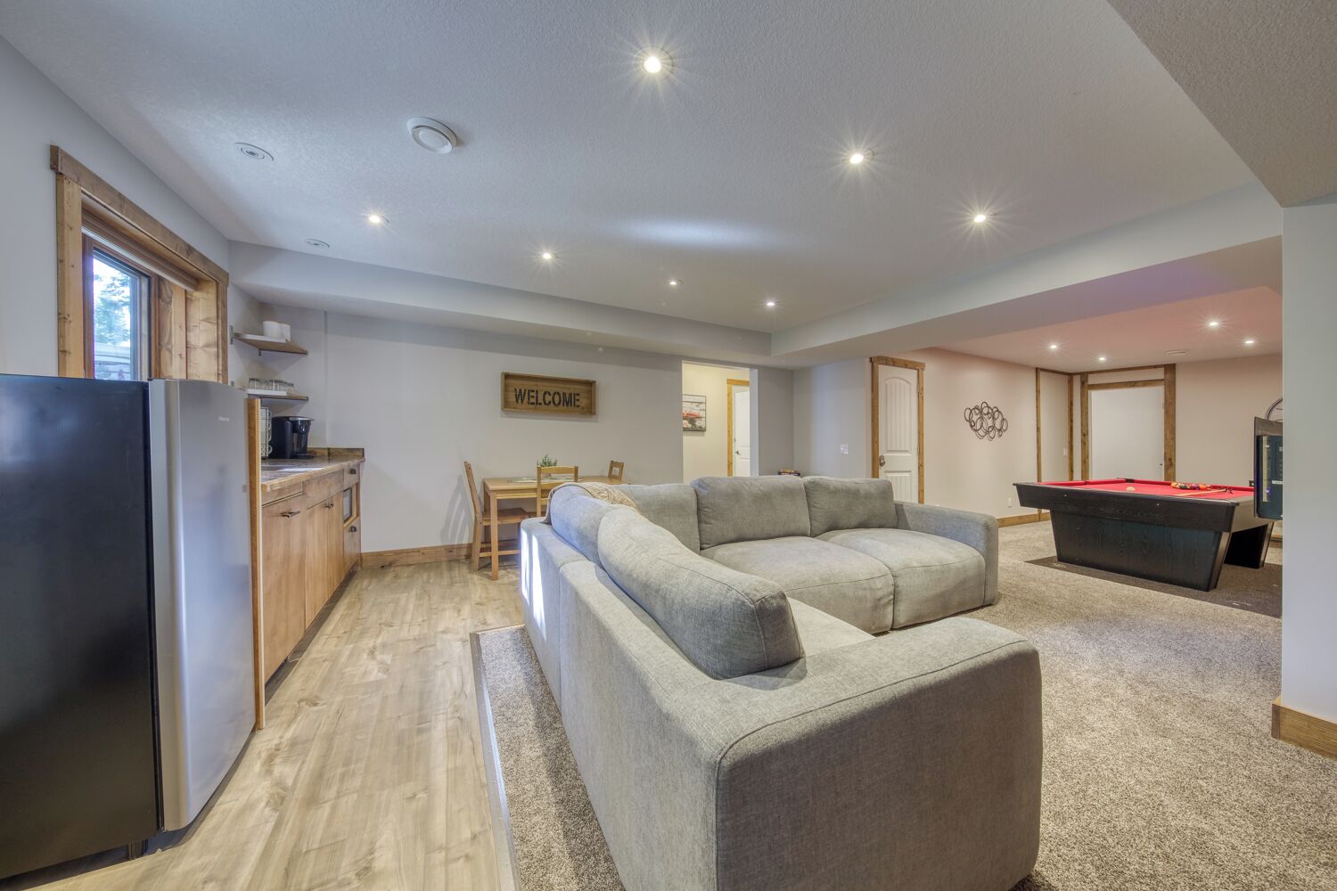 Een moderne woonkamer met een grijze modulaire bank, een pooltafel, een keuken met houten kasten, en een bord met de tekst 'WELCOME' aan de muur.