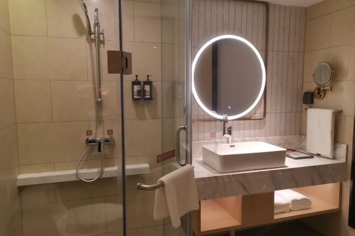 Un baño moderno con ducha de vidrio, espejo redondo iluminado, lavamanos blanco y un perchero para toallas.