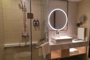 Un baño moderno con ducha de vidrio, espejo redondo iluminado, lavamanos blanco y un perchero para toallas.