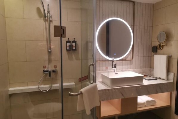 A modern bathroom with a glass-enclosed shower, a round mirror with a light, a white sink, and a towel rack.