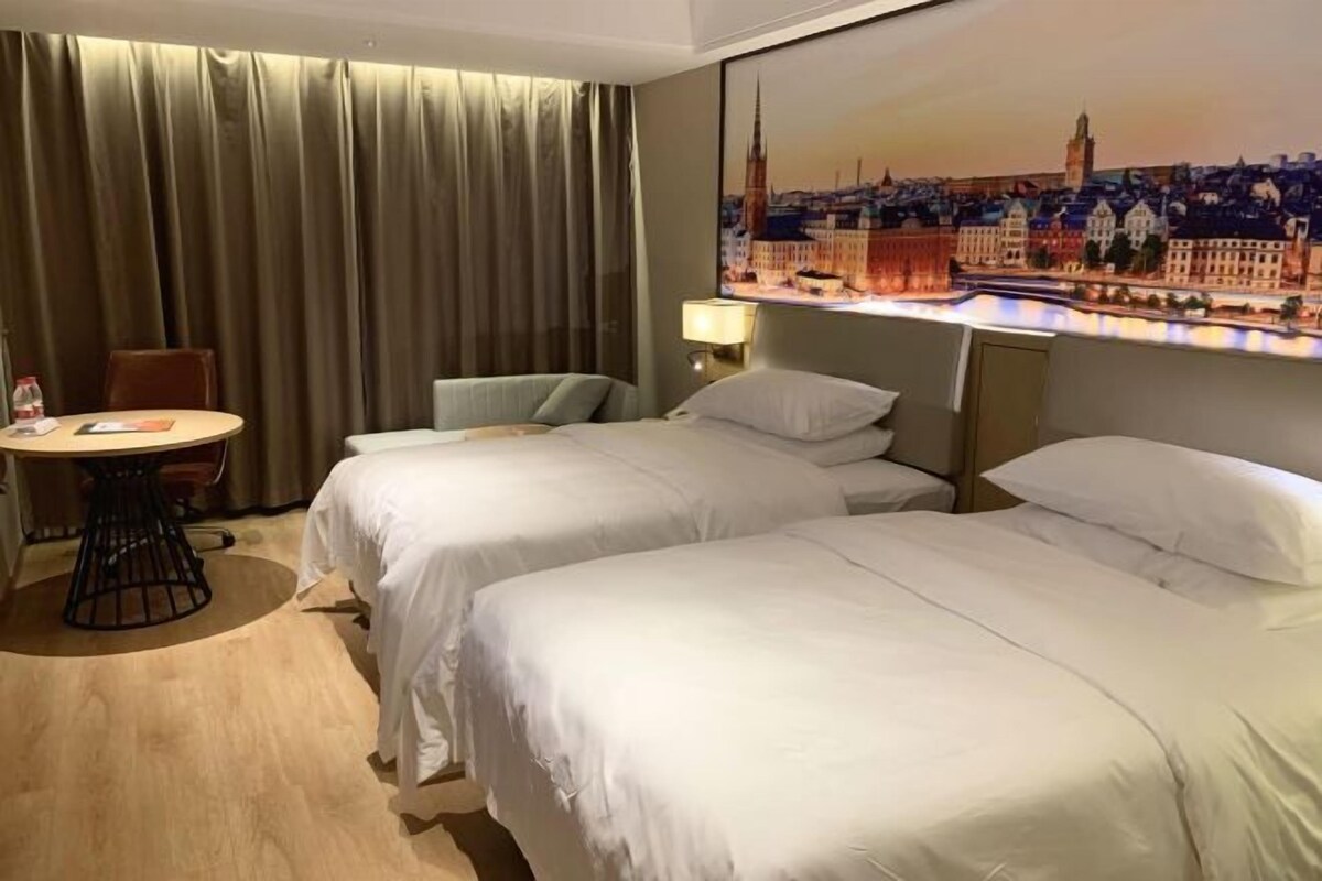 Una habitación de hotel con dos camas, una mesa, una silla, una lámpara y un cuadro de un paisaje urbano en la pared.